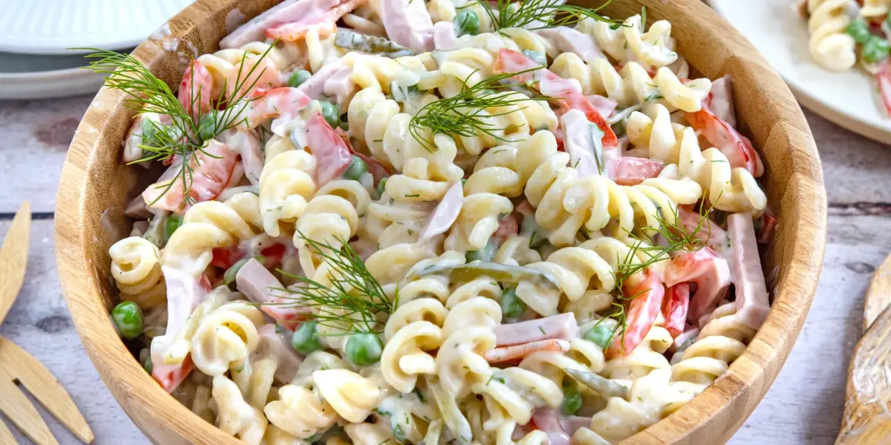 Ein großer Holzteller voller cremigen Nudelsalat mit Schinken, Erbsen und Paprika. Mit Dill garniert, sieht er aus wie der beste Nudelsalat!
