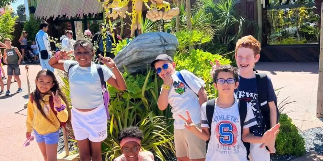 Des enfants joyeux font des signes de paix devant une attraction, un avis "supernova juniors" est visible.