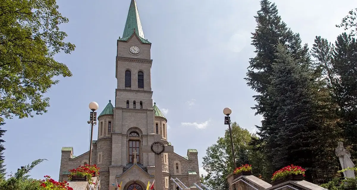 Słynny kościół w Zakopanem, z kamienną fasadą i zieloną wieżą, otoczony drzewami i ozdobiony czerwonymi kwiatami.