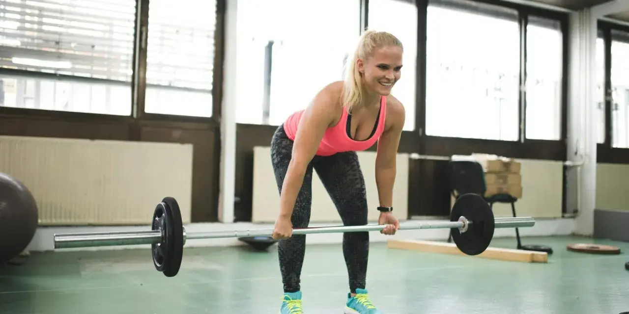 En kvinna lyfter skivstång i ett gym. Hon ler och ser fokuserad ut, en bra start på hur blir man stark.
