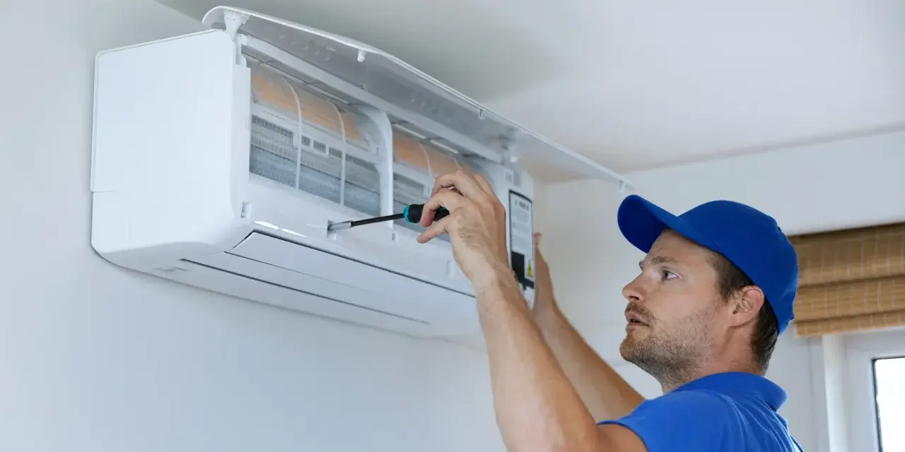 Un technicien en bleu utilise un tournevis pour ajuster un climatiseur mural, assurant le bon fonctionnement pour recharger clim maison.