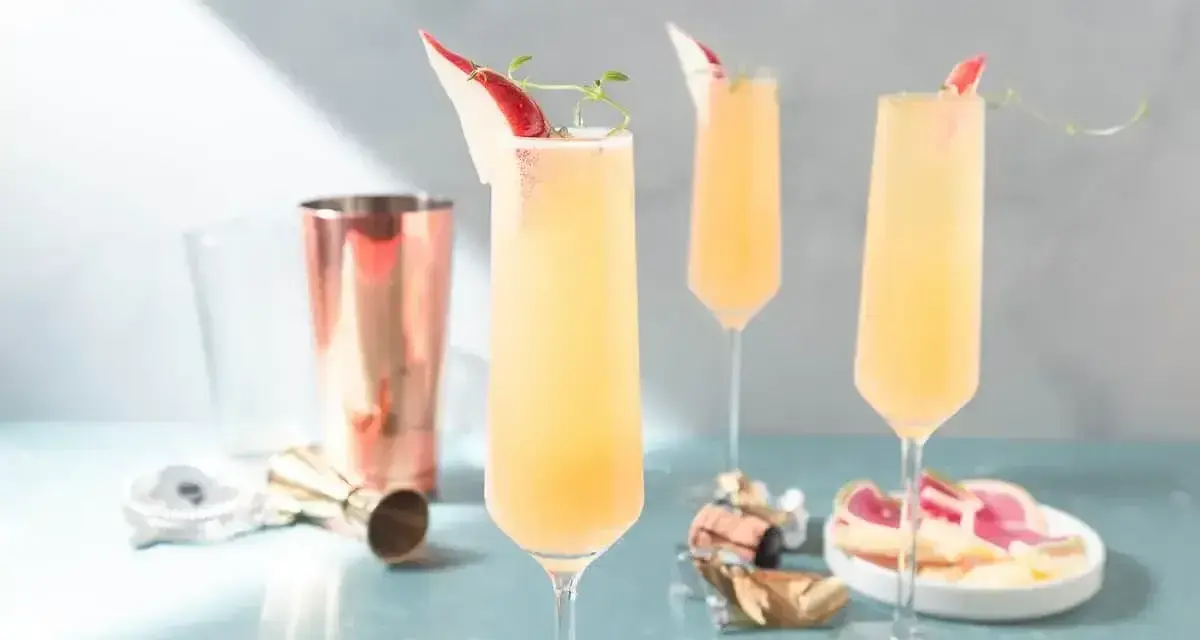 Tre eleganta glas med en ljusgul drink med gin och prosecco, garnerade med en tunn skiva röd frukt och en kvist.