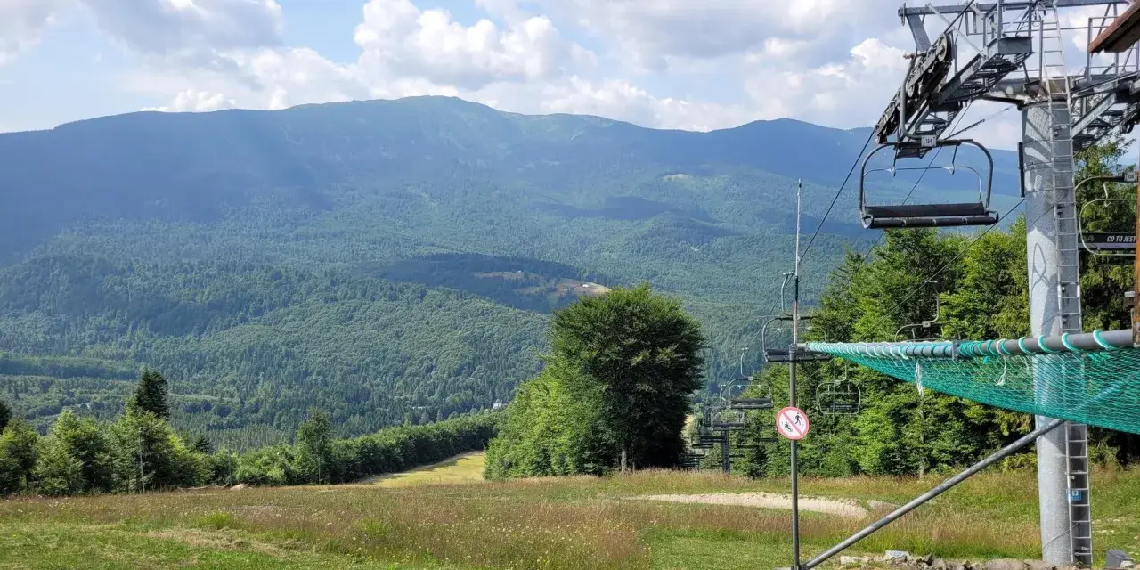 Widok na góry i lasy, z widoczną kolejką krzesełkową. W oddali majaczą kolejne pasma górskie, a na pierwszym planie trawiasty stok, który latem służy jako **zawoja szlaki**.