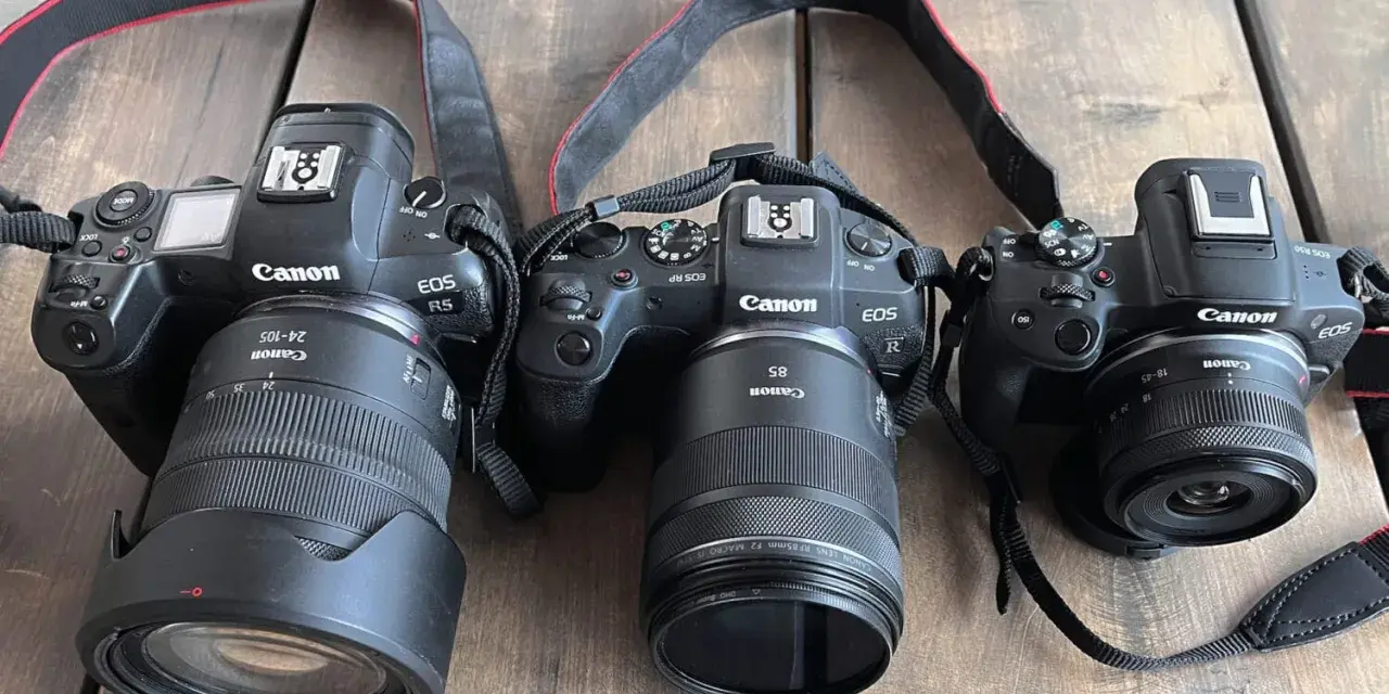 Trzy aparaty Canon EOS R, RP i R5 leżą obok siebie na drewnianym blacie. To **pełna klatka** sprzętu fotograficznego.