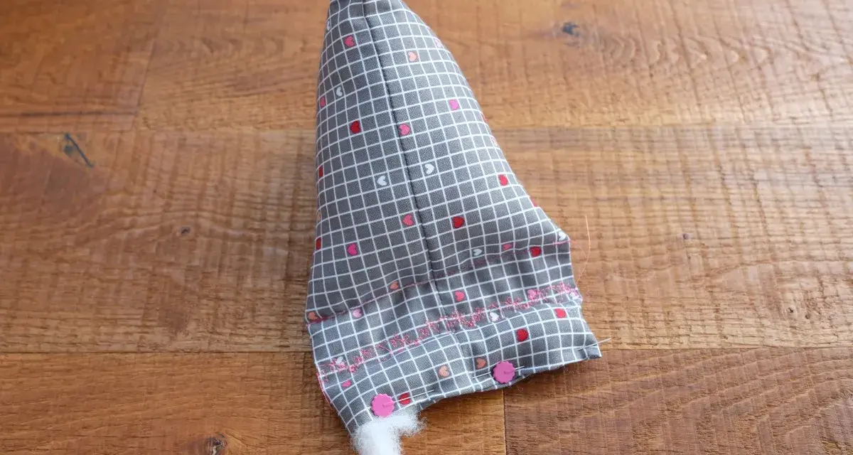 Un berlingot tissu gris à petits cœurs roses et blancs, en cours de rembourrage, est posé sur un parquet en bois.