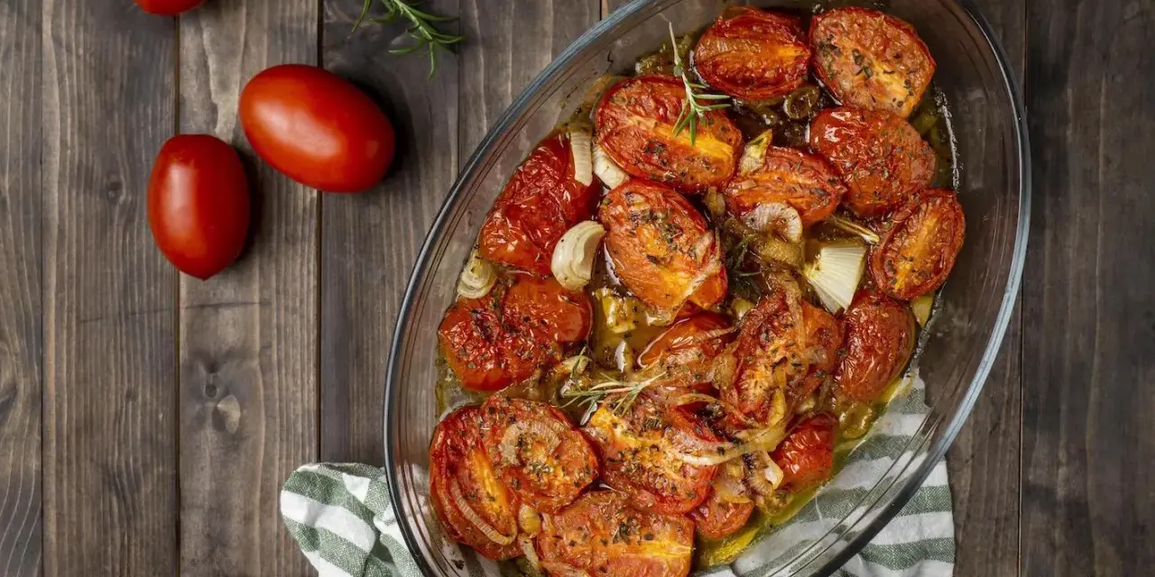 Pomodori gratinati al forno, dorati e succosi, con cipolle e rosmarino in una teglia di vetro su un tavolo di legno.
