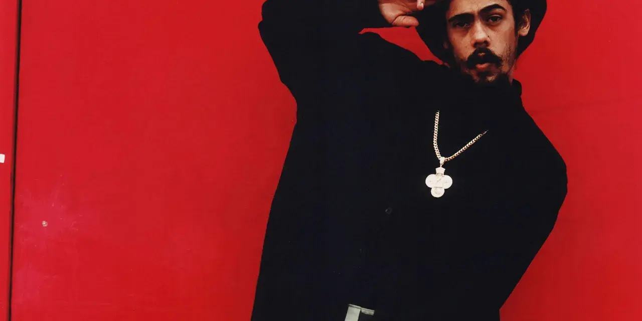 Mann mit Afro und Schnurrbart, schwarzer Rollkragenpullover, goldene Kette mit Kreuzanhänger, vor rotem Hintergrund. Er salutiert. "I shot the sheriff" könnte man sagen.