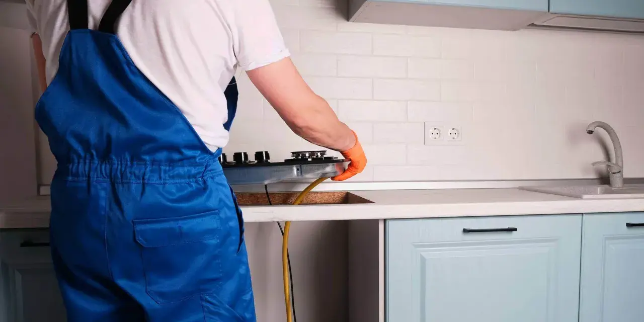 Instalator w niebieskim kombinezonie podłącza kuchenkę gazową w kuchni.
