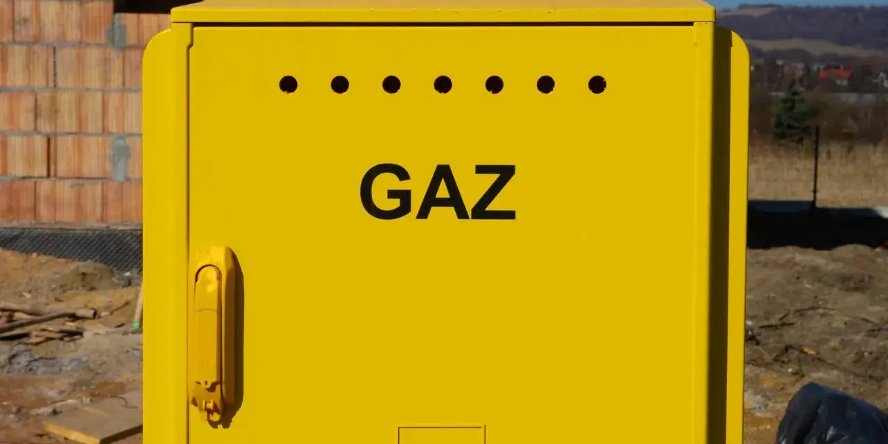 Żółta szafka gazowa z napisem "GAZ" i otworami wentylacyjnymi. Obok budowa domu, w tle krajobraz. Przyłącze gazowe krok po kroku.