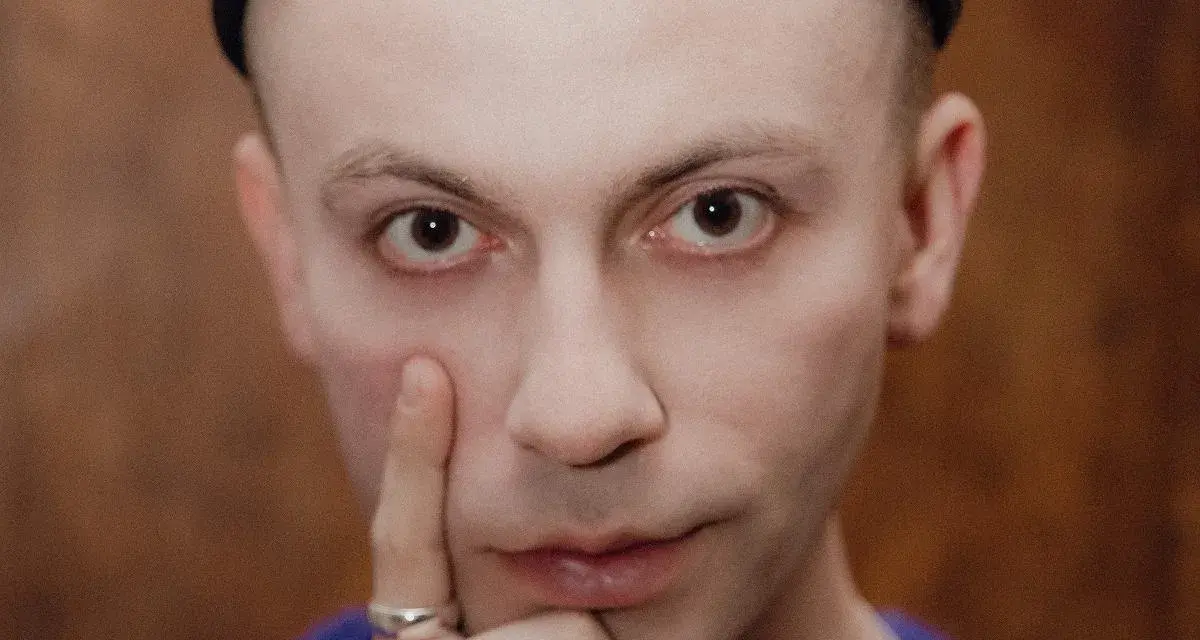 Zbliżenie na twarz Arka Kłusowskiego w czarnej czapce, z palcem przy policzku.