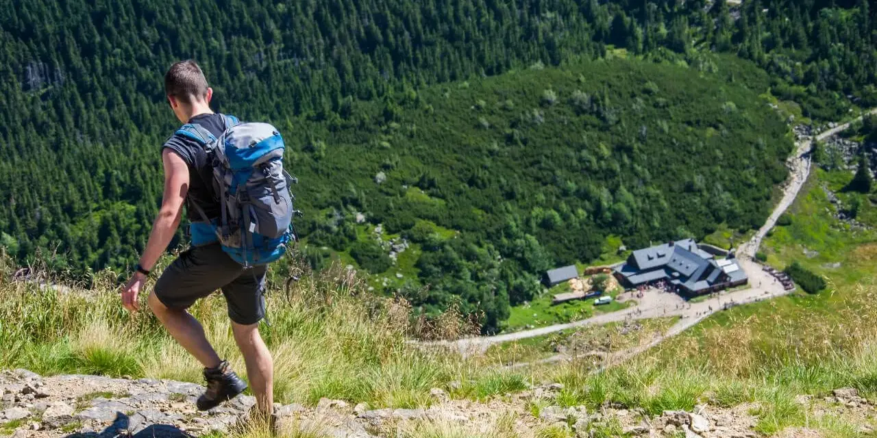 Mężczyzna z plecakiem wędruje po kamienistym szlaku w Karkonoszach, podziwiając widok na schronisko i las.