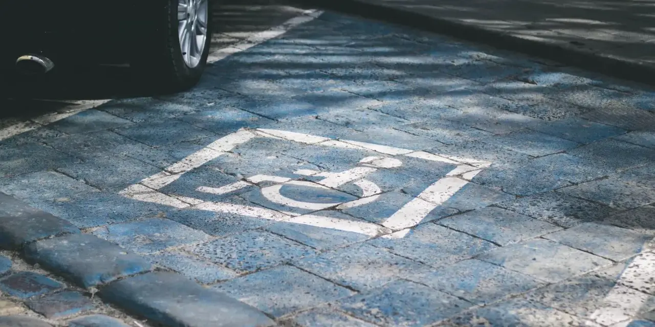 Niebieskie miejsce parkingowe dla niepełnosprawnych pod blokiem, z symbolem wózka inwalidzkiego. Obok widoczna część czarnego samochodu.