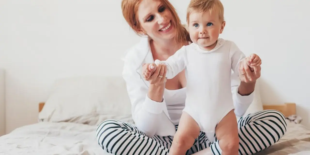 Uśmiechnięta mama pomaga swojemu 8 miesięcznemu dziecku w pierwszych krokach na łóżku.
