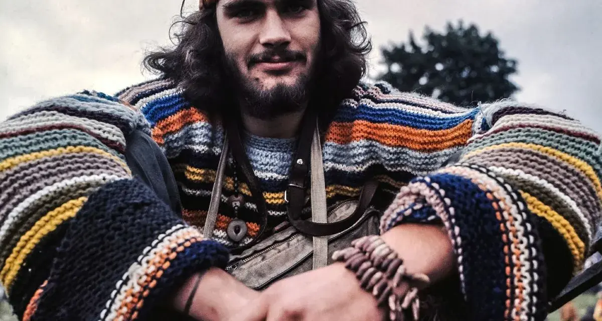 Młody mężczyzna w kolorowym swetrze w paski i opasce na głowie, uśmiecha się. Wygląda jak prawdziwy hippie.