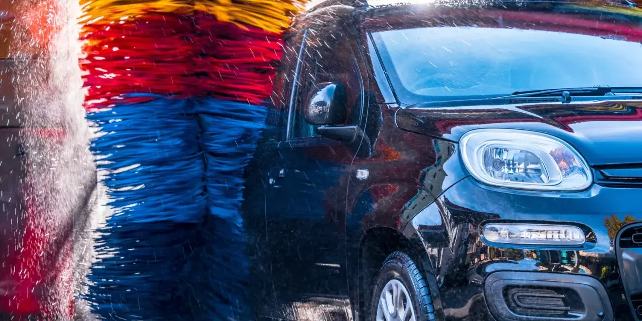 Czarna, błyszcząca karoseria samochodu w myjni automatycznej, otoczona kolorowymi szczotkami. Czy myjnia automatyczna niszczy lakier? Woda spływa po aucie.