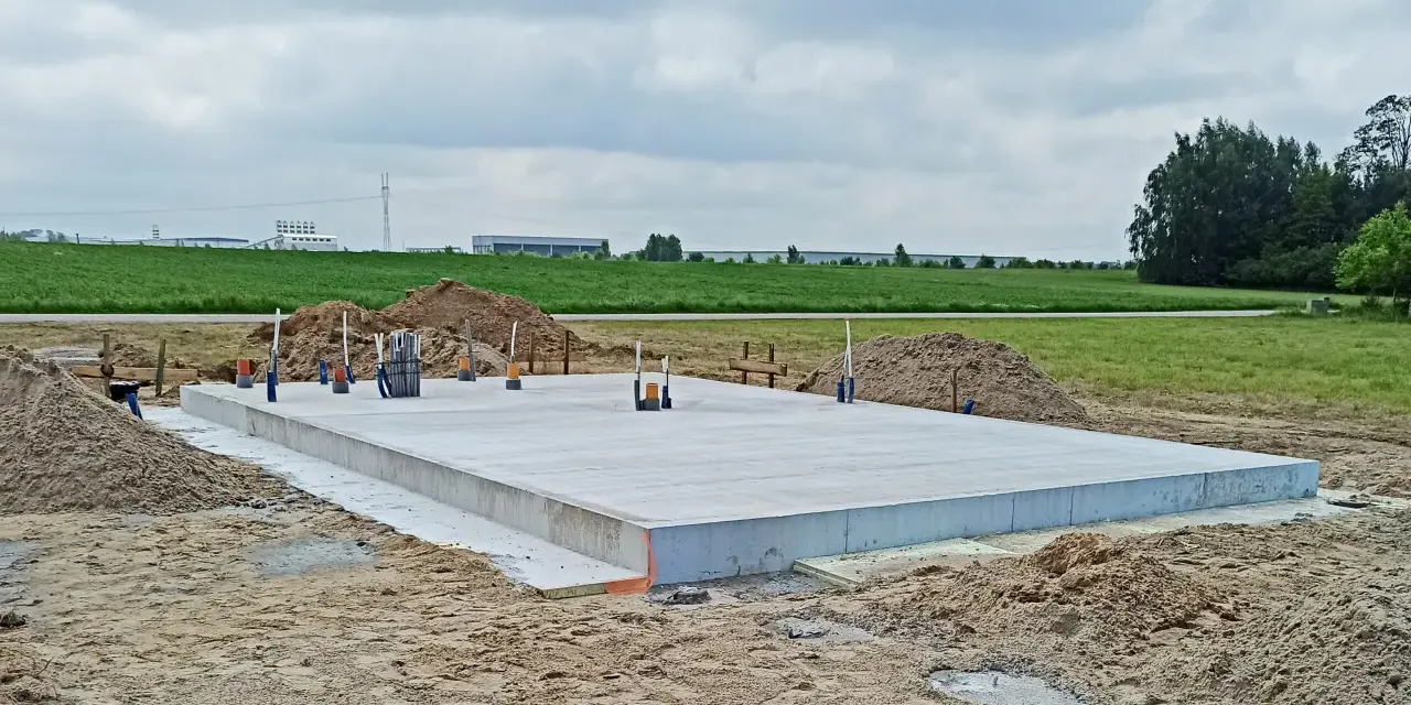 Solidna płyta fundamentowa, gotowa na budowę. Wokół piasek i zielone pola.