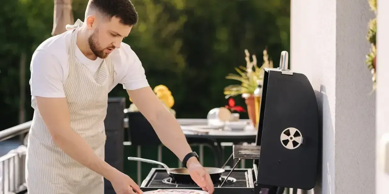 Mężczyzna w fartuchu przygotowuje mięso na grillu na balkonie. Czy można robić grilla na balkonie? Tak, jeśli jest bezpiecznie!