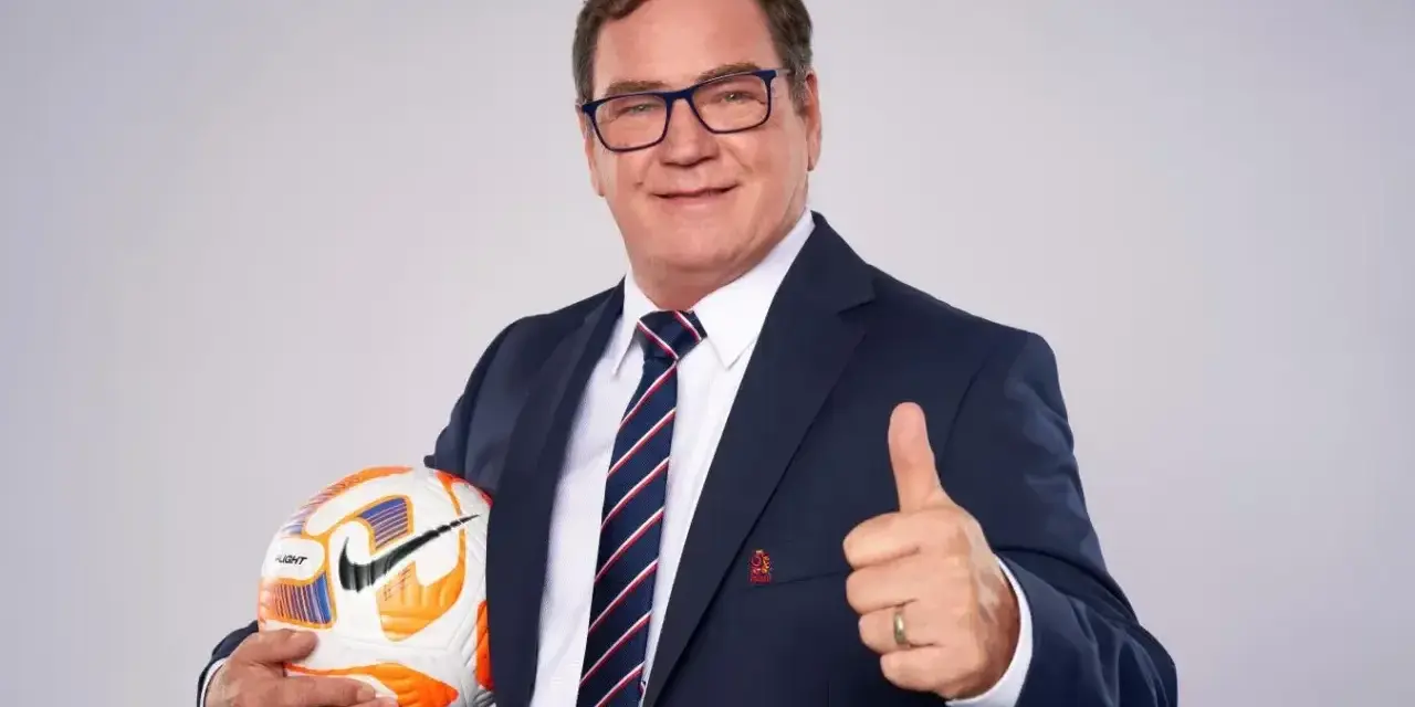 Uśmiechnięty trener reprezentacji Polski w garniturze trzyma piłkę i pokazuje kciuk w górę.
