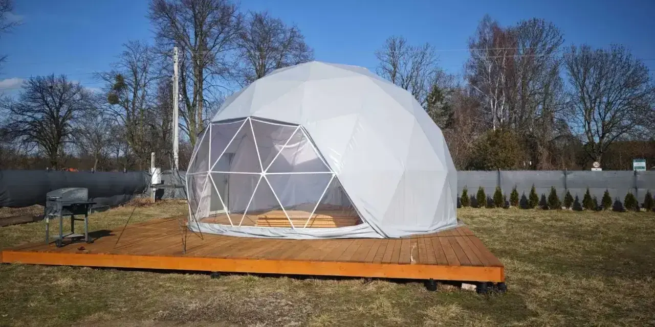 Nowoczesny glamping nad jeziorem. Biała kopuła na drewnianym tarasie, obok grill. Idealne miejsce na relaks.