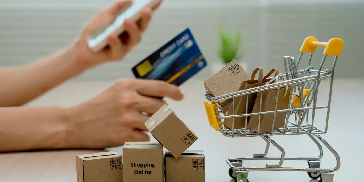 Manos sostienen un teléfono y tarjeta de crédito, junto a un carrito de compras con cajas y bolsas. El modelo 369 representa la facilidad de comprar en línea.