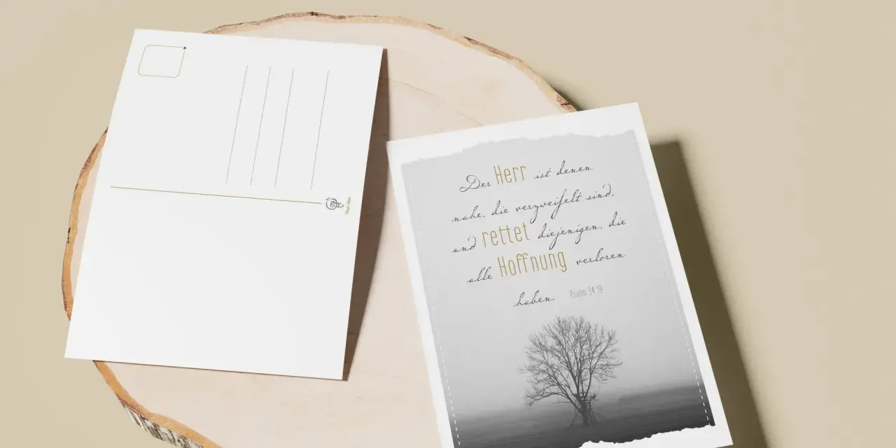 Zwei Postkarten liegen auf einem Holzstamm. Eine zeigt einen einsamen Baum im Nebel und den Spruch: "Der Herr ist denen nahe, die verzweifelt sind und rettet diejenigen, die alle Hoffnung verloren haben.
