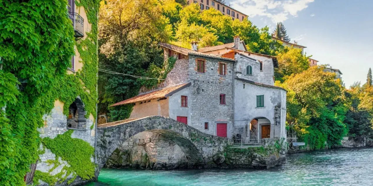 Antica casa in pietra con edera su un lago turchese, un ponte ad arco e colline verdi. Un luogo incantevole per i cammini in Lombardia.