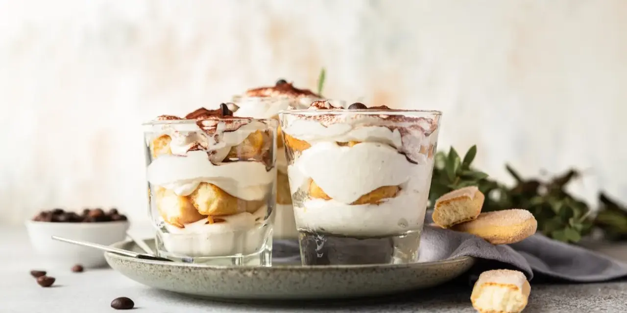 Pyszne desery włoskie: tiramisu w szklankach, udekorowane kakao i kawą. Obok kawałki biszkoptów i ziarna kawy.