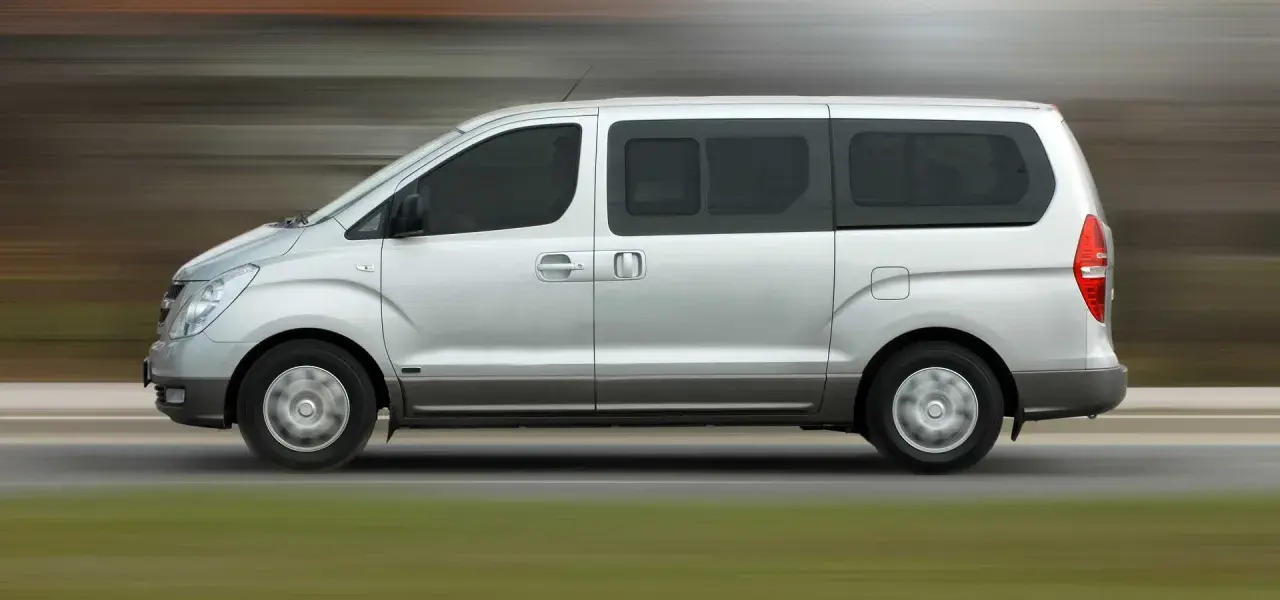 Un mini bus argenté roule sur une route. Ce véhicule spacieux est idéal pour les familles ou les groupes, et peut être conduit avec un permis B.