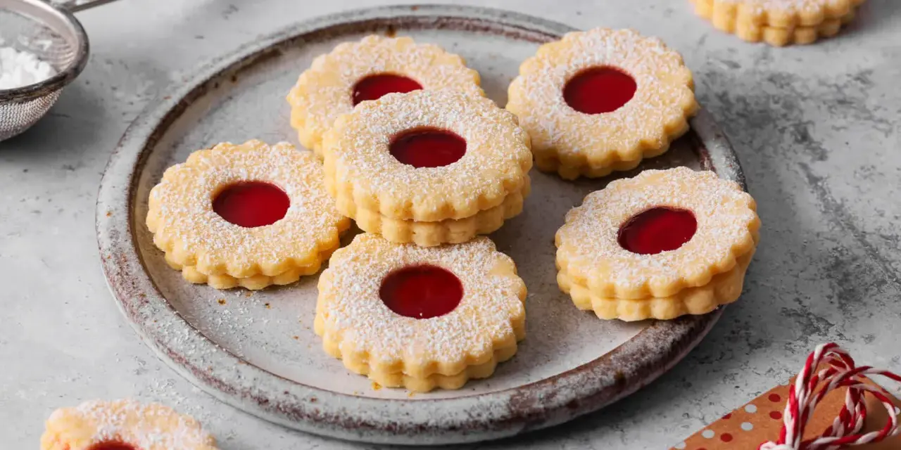 Ein Teller voller **die besten Spitzbuben der Welt**, bestäubt mit Puderzucker, mit leuchtend roter Marmeladenfüllung.