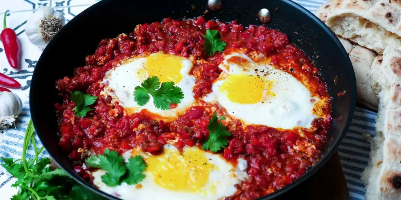 Herzhaftes Shakshuka-Rezept in einer Pfanne mit pochierten Eiern in würziger Tomatensoße, garniert mit Koriander. Dazu gibt es Fladenbrot.