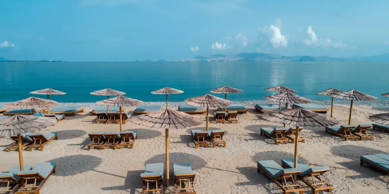 Plaża z leżakami i parasolami, idealna na relaks nad morzem. W oddali widać góry i błękitne niebo.