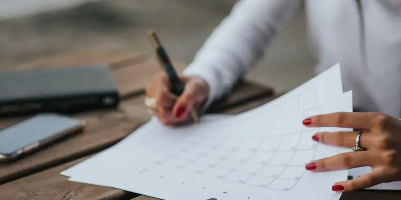 Une main avec des ongles rouges écrit sur un calendrier, planifiant peut-être la date de mariage. Un téléphone et un carnet sont à proximité.