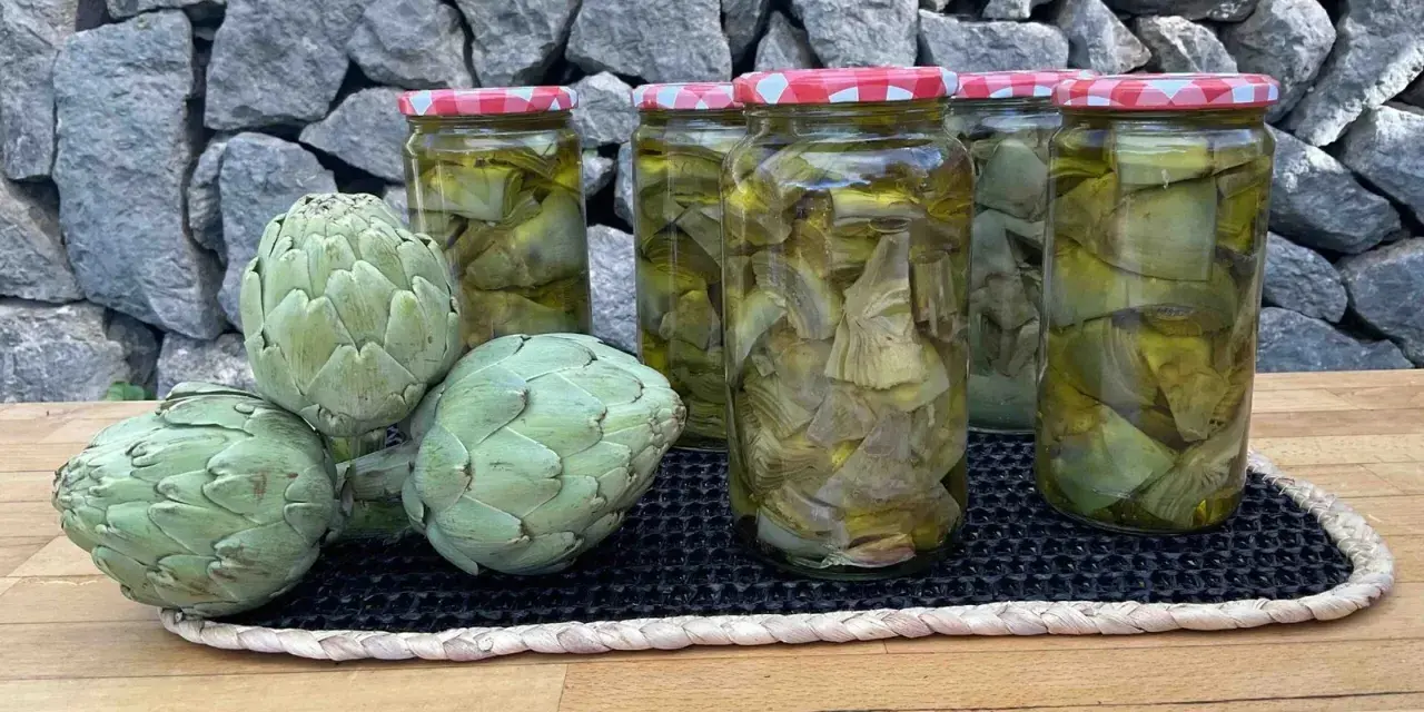 Alcachofas frescas y en conserva en frascos, sobre una mesa de madera y fondo de piedra.