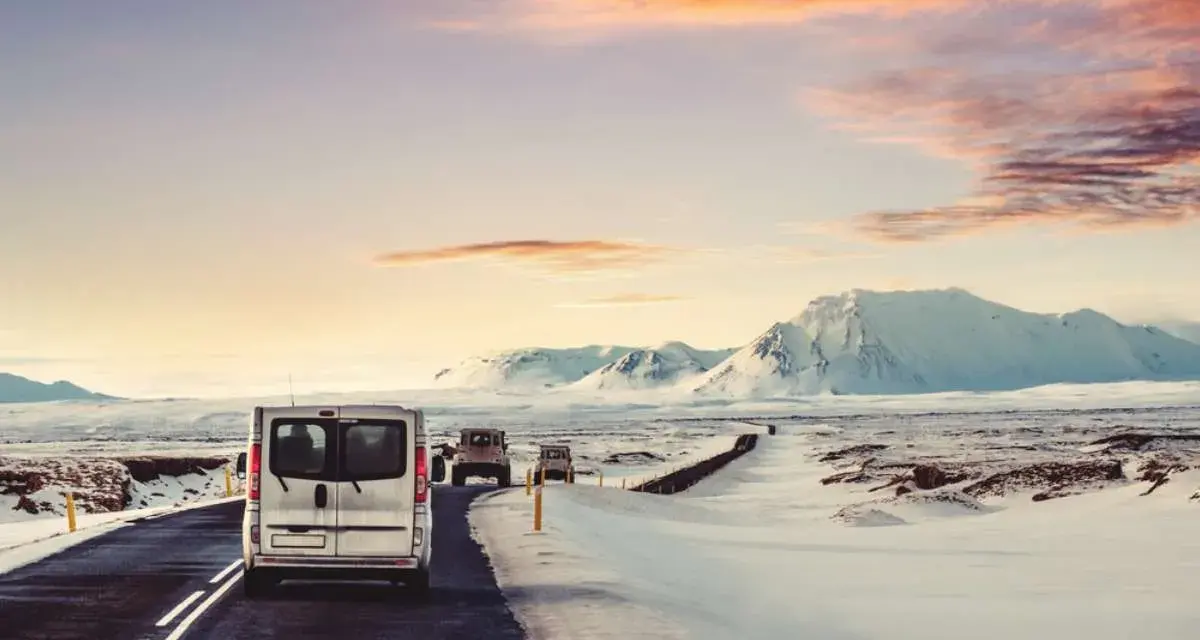 A silver van leads a convoy of vehicles driving in Iceland in December, past snow-covered mountains under a dramatic sunset sky.
