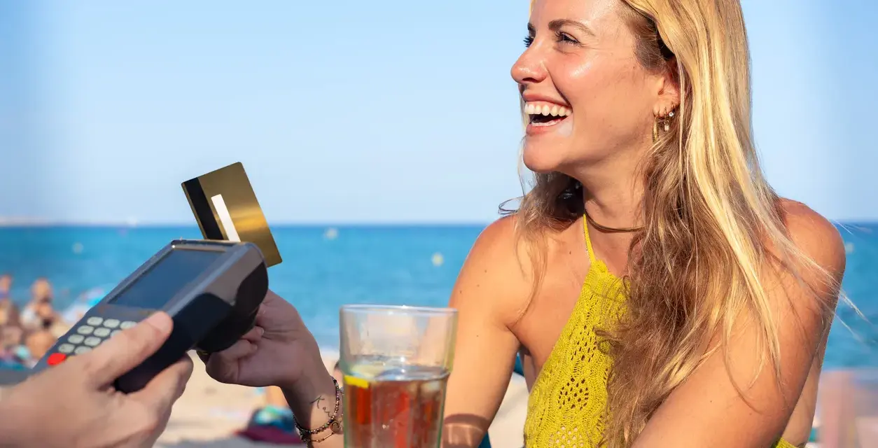Lachende Frau bezahlt am Strand mit Karte. Selbst mit Krankengeld ist Urlaubsgeld drin!