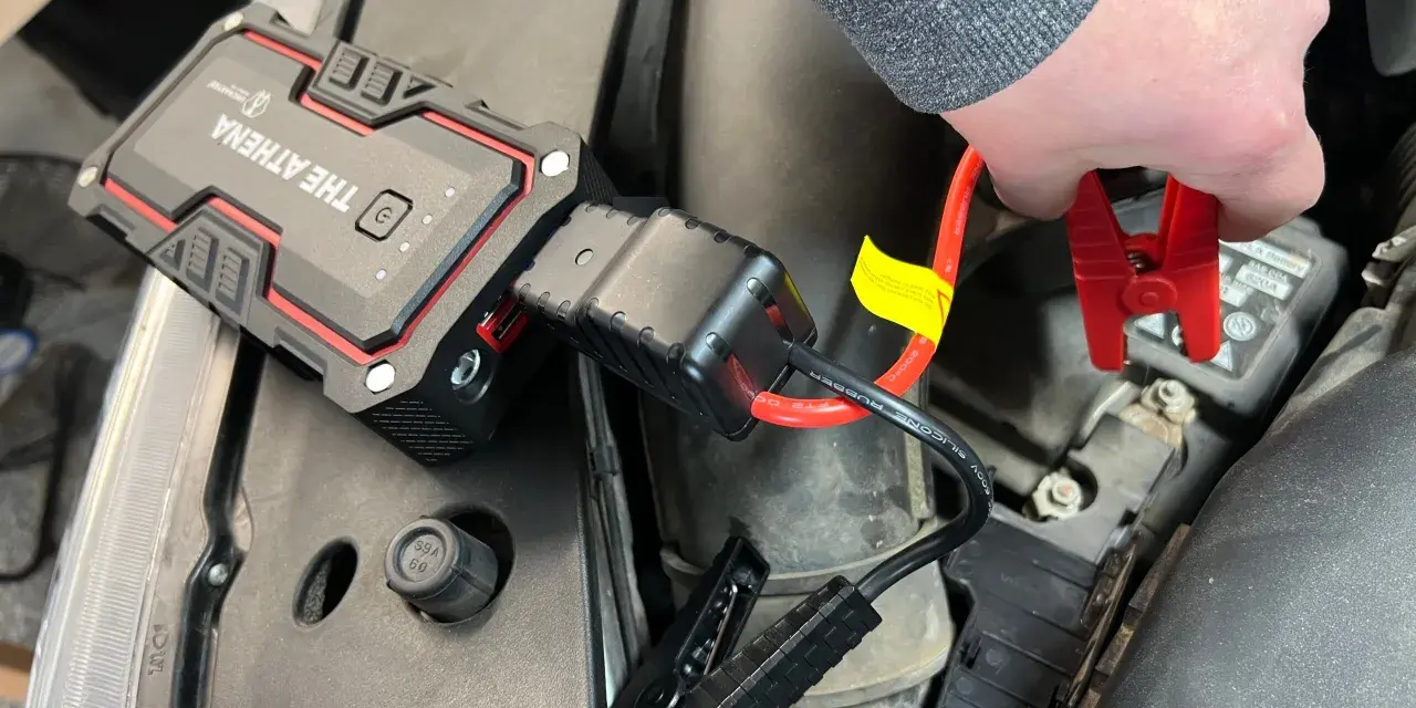 A hand connects red jumper cables from "The Athena" lithium jump starter to a car battery, demonstrating how lithium jump starters work by providing a temporary power boost.