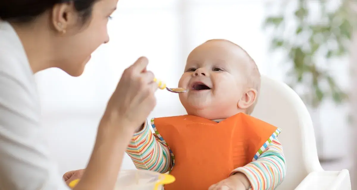 Mama karmi uśmiechniętego niemowlaka łyżeczką. To idealny przykład, co na śniadanie dla niemowlaka – zdrowo i radośnie!