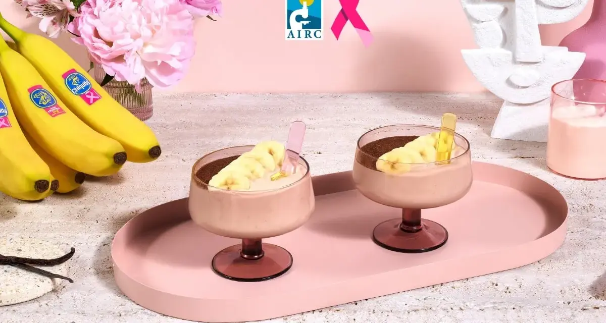 Due coppe di cremoso gelato alla banana, guarnite con fettine di banana e cacao, su un vassoio rosa. Banane fresche e fiori rosa sullo sfondo.