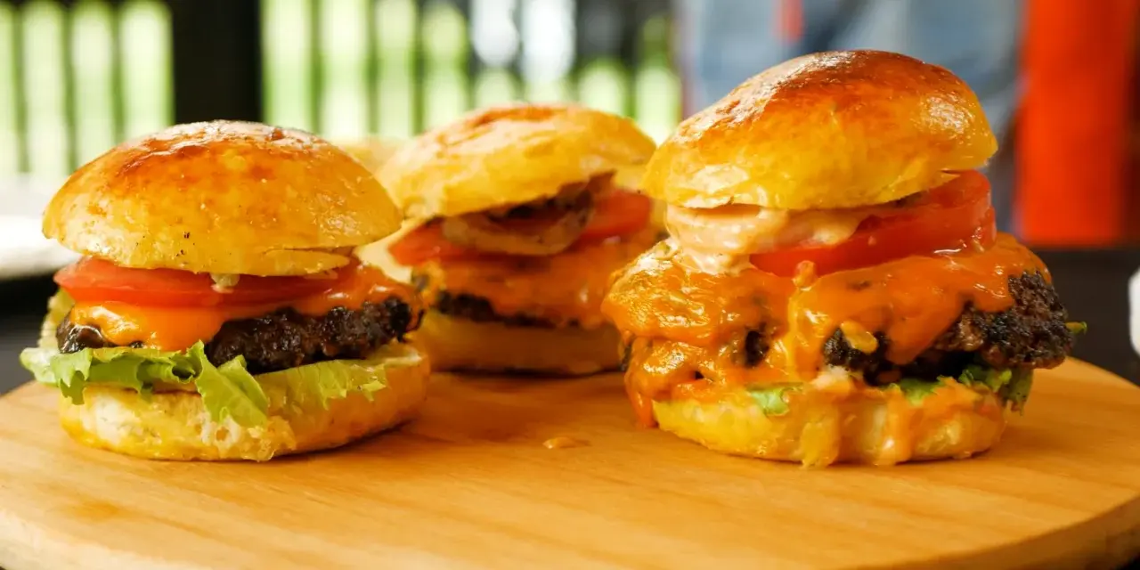 Trzy soczyste burgery z roztopionym serem, pomidorem i sałatą na desce.