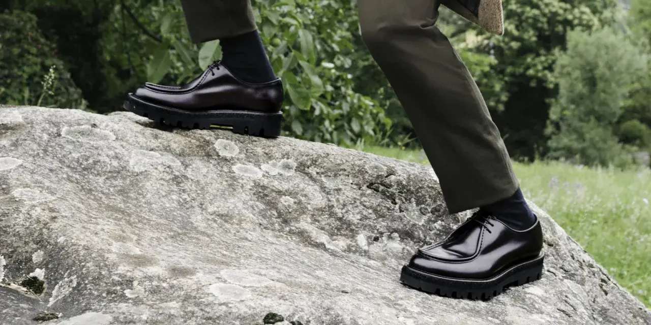Scarpe oxford derby lucide su una roccia, con pantaloni verdi e sfondo verdeggiante.