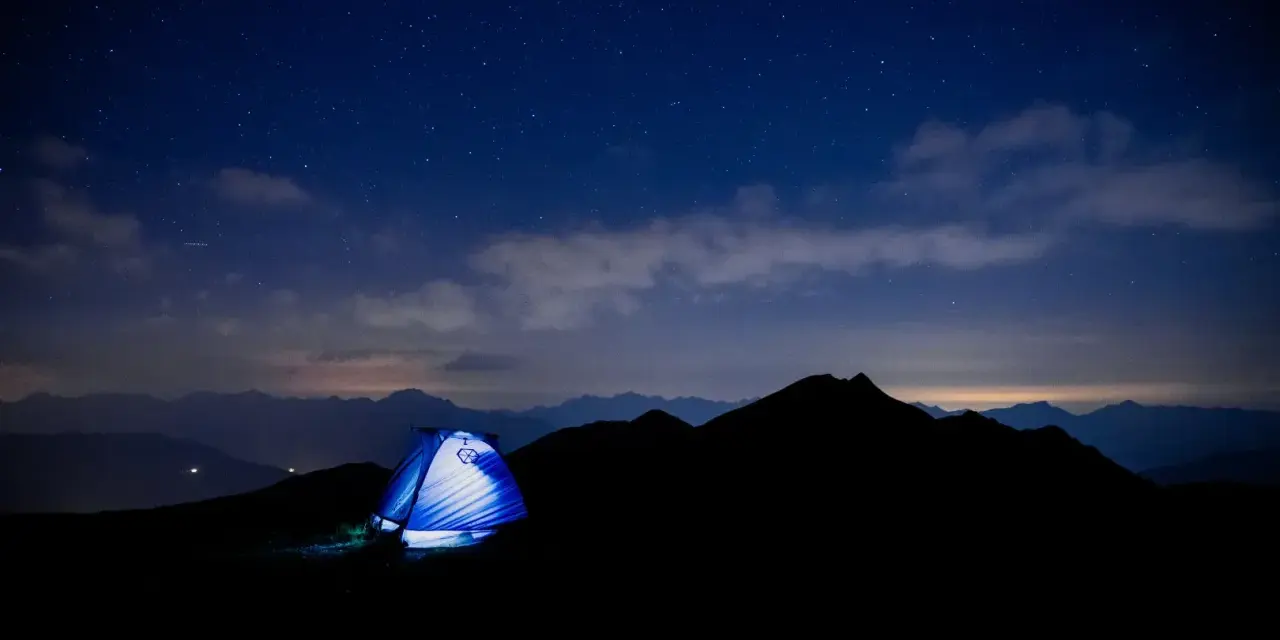 Une tente bleue s'illumine sous un ciel étoilé, témoin silencieux d'un trek inoubliable au cœur des montagnes.