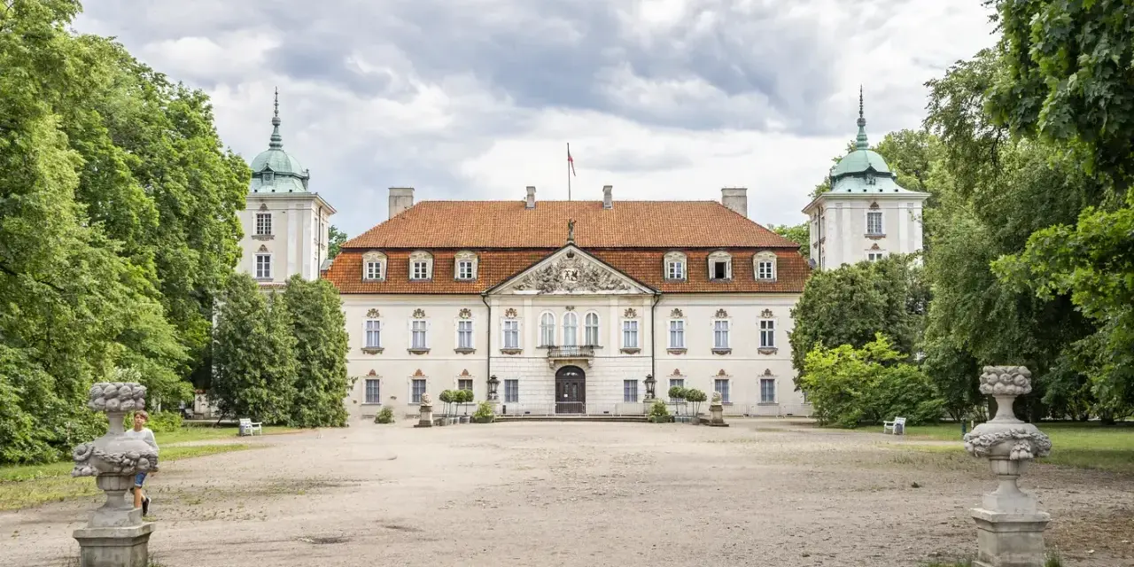 Pałac w Nieborowie, biała rezydencja z czerwonym dachem, otoczona zielenią. Dwie wieżyczki z zielonymi kopułami po bokach.
