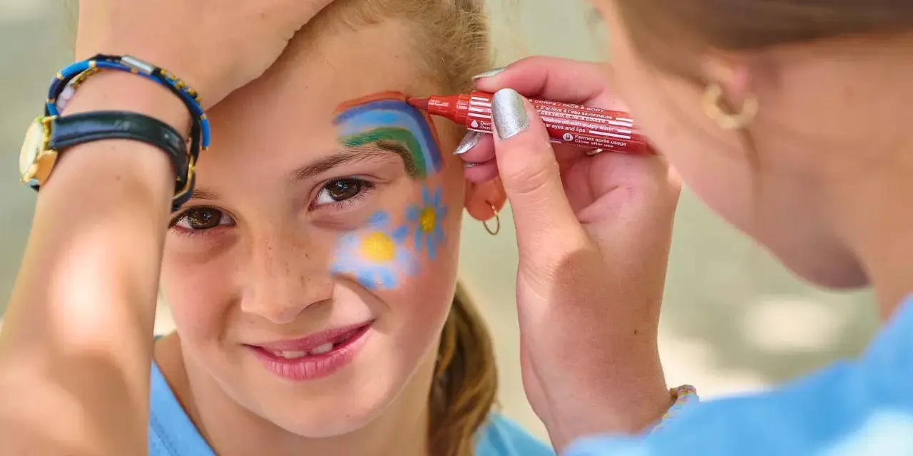 Une petite fille souriante se fait maquiller sur la joue un arc-en-ciel et des fleurs, au stand maquillage d'une kermesse.