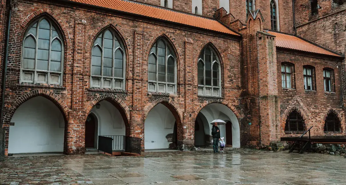 Zamek w Kwidzynie, z ceglanymi arkadami i gotyckimi oknami, skrywa tajemnice. Dwoje ludzi z parasolem spaceruje po mokrym dziedzińcu.