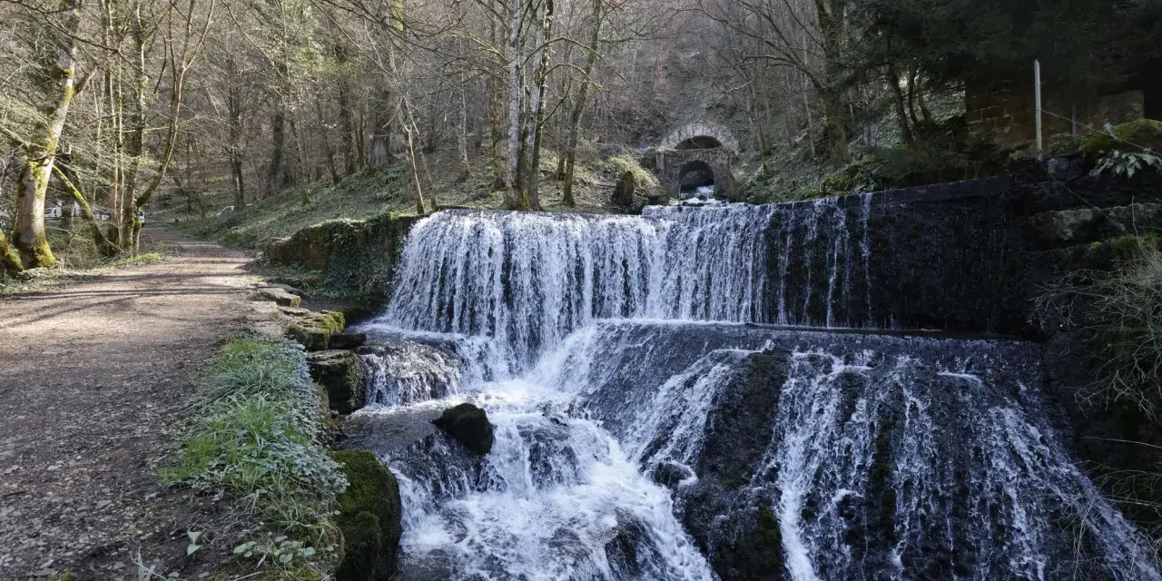Cascade d'eau dans une forêt, un chemin de terre longe la rivière. Au fond, un tunnel voûté suggère une source d'arcier.