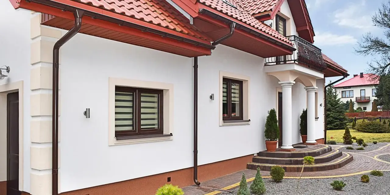 Elegancki dom z białym tynkiem i brązowymi detalami. Cokół wykończony ozdobnym tynkiem mozaikowym dodaje mu charakteru.