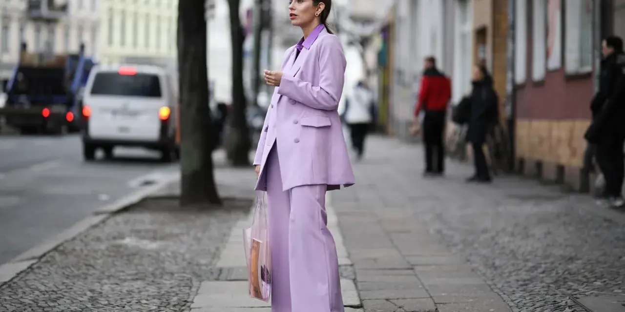 Elegancka kobieta w garniturze w kolorze liliowym, z transparentną torbą, idzie ulicą.