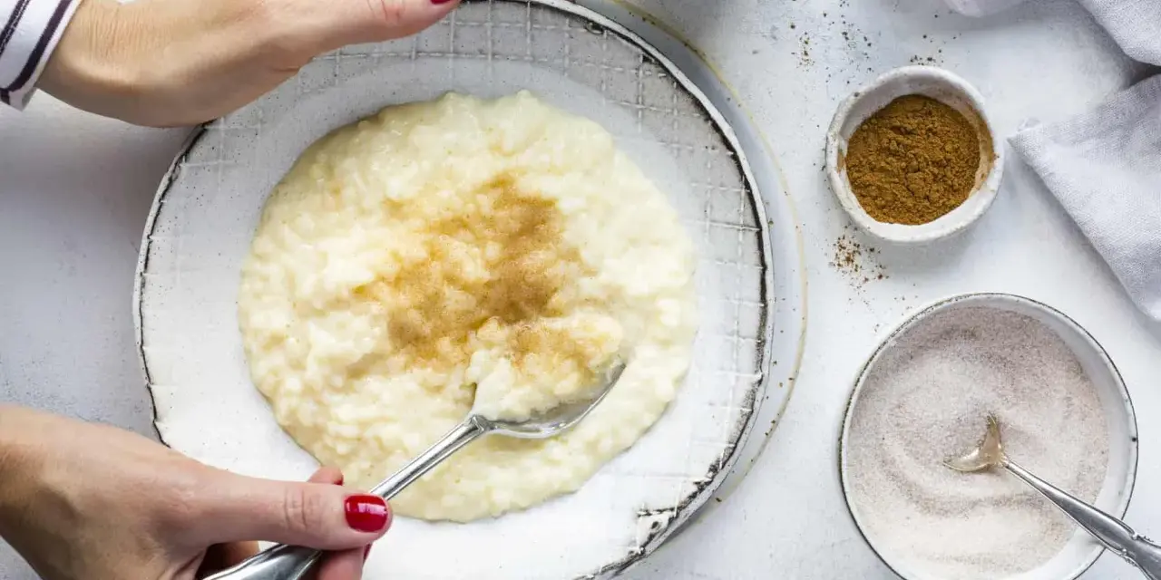 Cremiger Milchreis mit Vanillepudding, bestreut mit Zimt. Eine Hand mit roten Nägeln hält einen Löffel bereit.