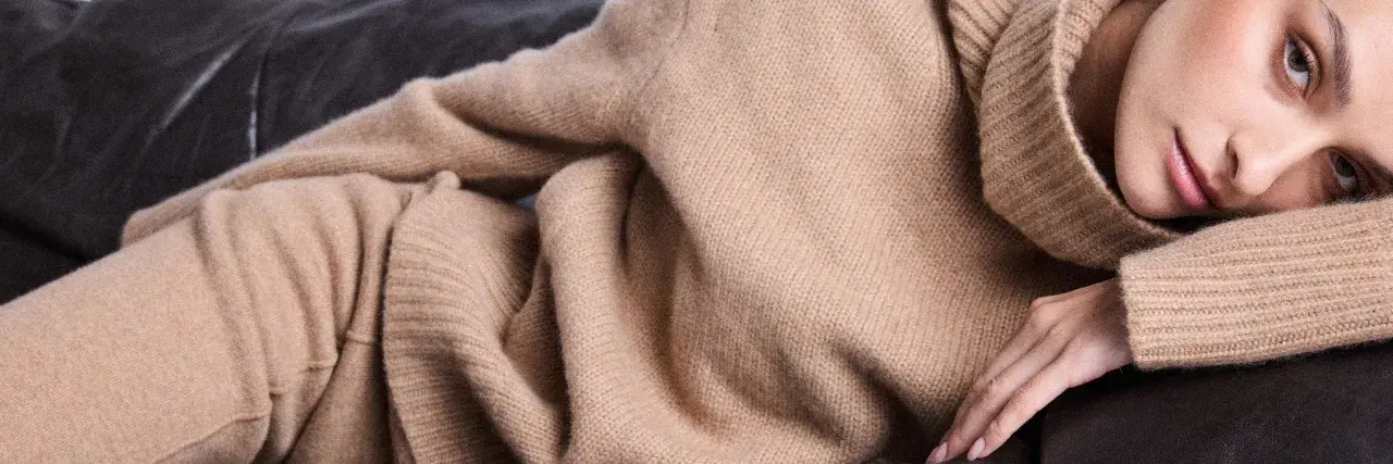 Eine Frau in einem beigen Kaschmirpullover liegt entspannt auf einem dunklen Sofa. Das Kaschmir waschen ist wichtig für die Pflege.