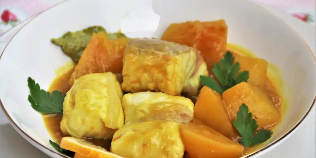Sabroso cazon en amarillo, con trozos de pescado blanco y patatas en una salsa dorada, decorado con perejil y rodajas de limón.
